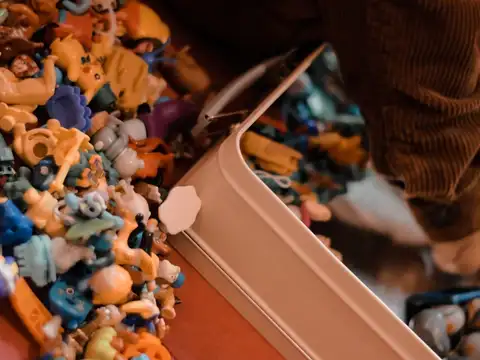 A large spread of small plastic collectible figures beside an open tin, with a seated person just visible behind them.