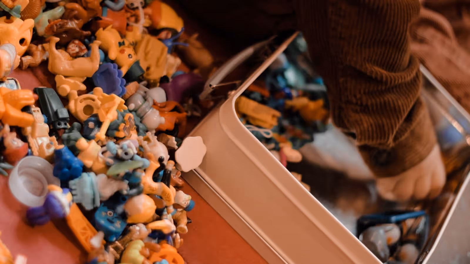 A large spread of small plastic collectible figures beside an open tin, with a seated person just visible behind them.