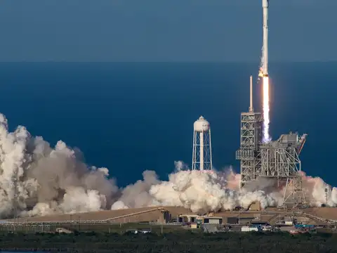 A white rocket rising from a coastal launch pad on a long orange exhaust flame, with a vast white plume of smoke rolling out across the ground and open blue sea filling the background.