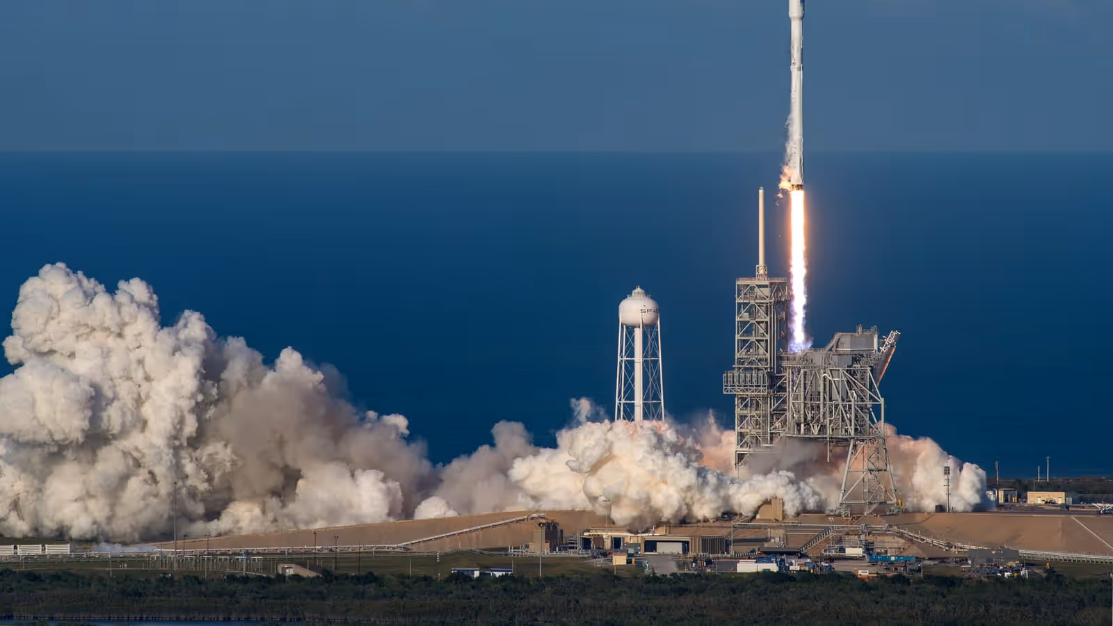 A white rocket rising from a coastal launch pad on a long orange exhaust flame, with a vast white plume of smoke rolling out across the ground and open blue sea filling the background.