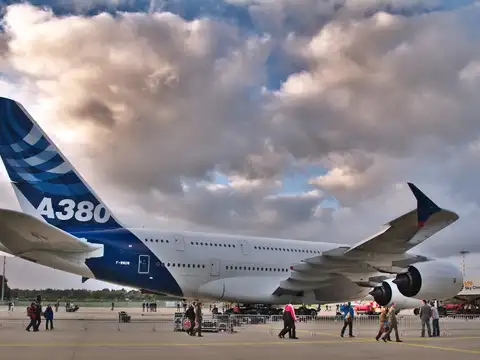 An Airbus A380 in blue Airbus livery parked on an airport apron under a cloudy sky, A380 lettered large on its tail fin, with people walking behind barriers in the foreground.