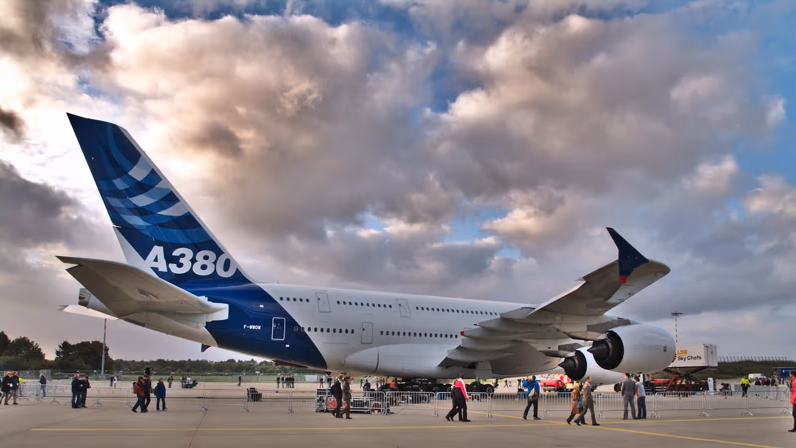An Airbus A380 in blue Airbus livery parked on an airport apron under a cloudy sky, A380 lettered large on its tail fin, with people walking behind barriers in the foreground.