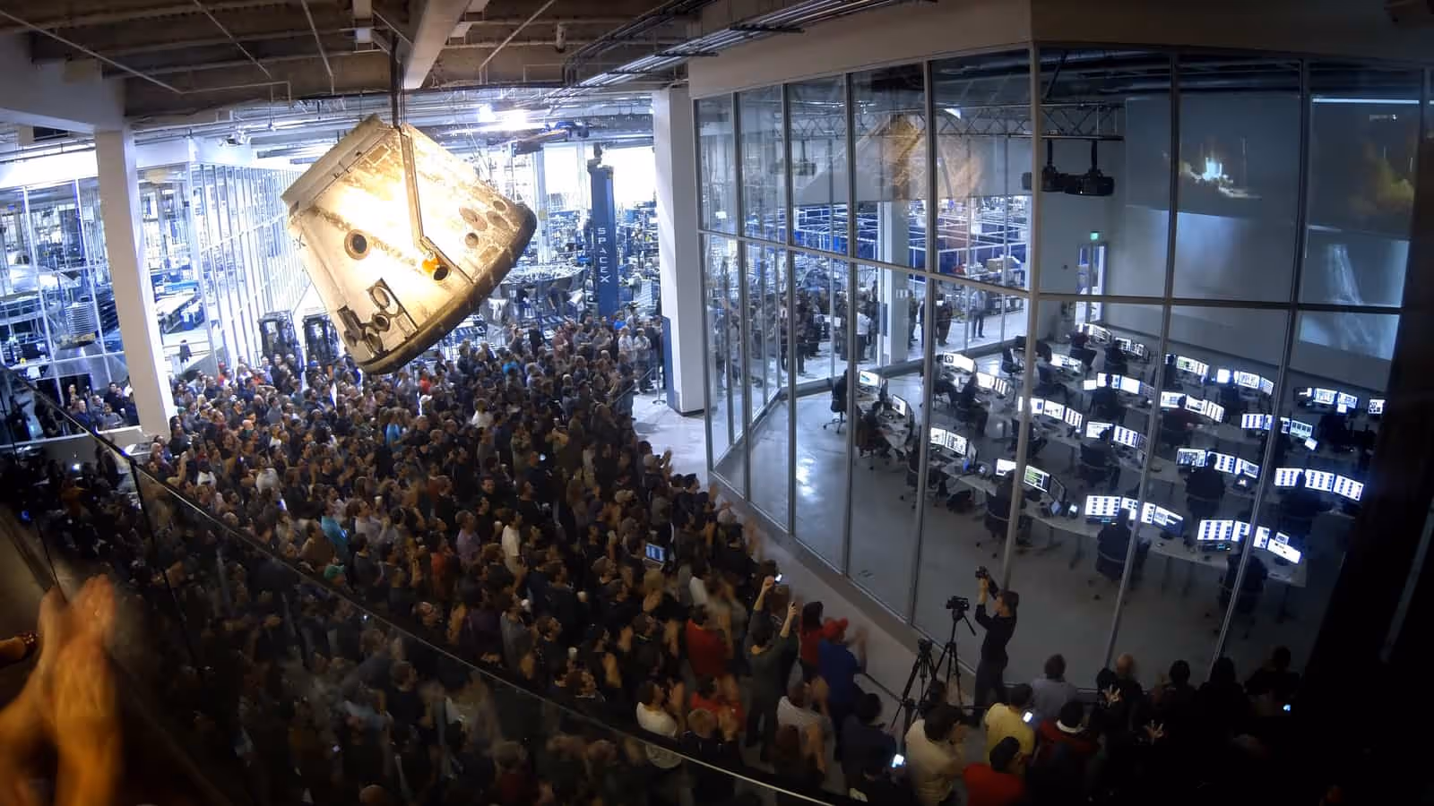 A large crowd standing on a factory floor with hands raised and phones held up, a scorched space capsule suspended from the ceiling above them, and a glass-walled control room full of monitors alongside.