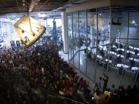 A large crowd standing on a factory floor with hands raised and phones held up, a scorched space capsule suspended from the ceiling above them, and a glass-walled control room full of monitors alongside.