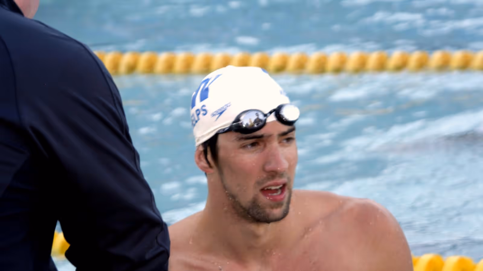 Michael Phelps at the side of a swimming pool in a white swim cap with goggles pushed up on his forehead, looking to one side, with lane ropes behind him.