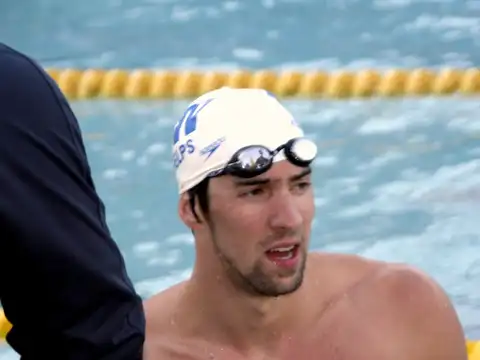 Michael Phelps at the side of a swimming pool in a white swim cap with goggles pushed up on his forehead, looking to one side, with lane ropes behind him.