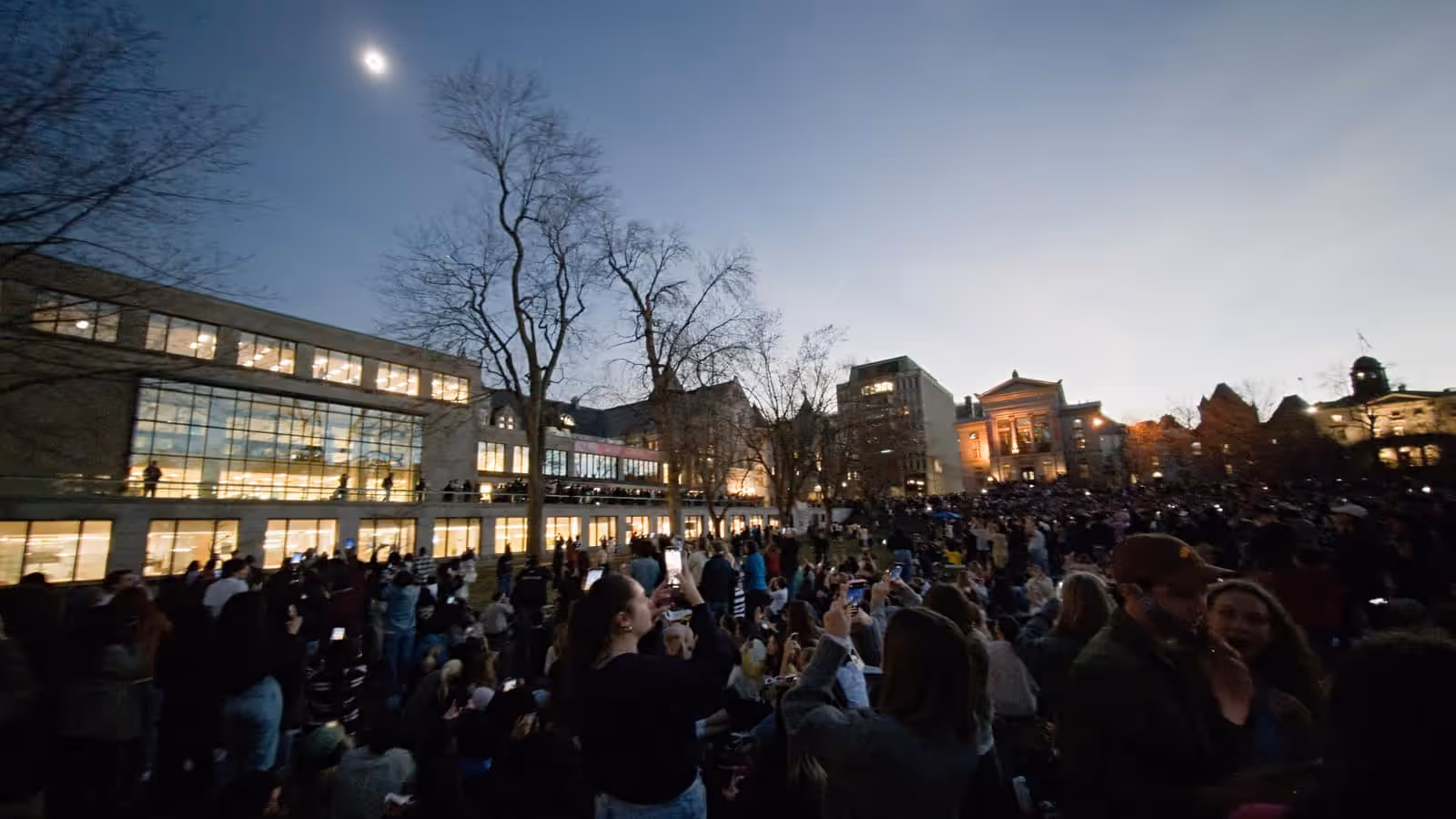 A large crowd standing on a campus lawn at dusk, many holding phones up towards a total solar eclipse showing as a bright ring high in the darkened sky above lit university buildings and bare trees.