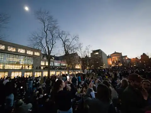 A large crowd standing on a campus lawn at dusk, many holding phones up towards a total solar eclipse showing as a bright ring high in the darkened sky above lit university buildings and bare trees.