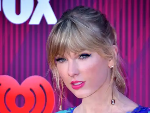 Taylor Swift facing the camera in a head-and-shoulders portrait, blonde hair up with a fringe, gold drop earrings and a blue sequinned top, in front of a magenta backdrop carrying the FOX and iHeartRadio logos.
