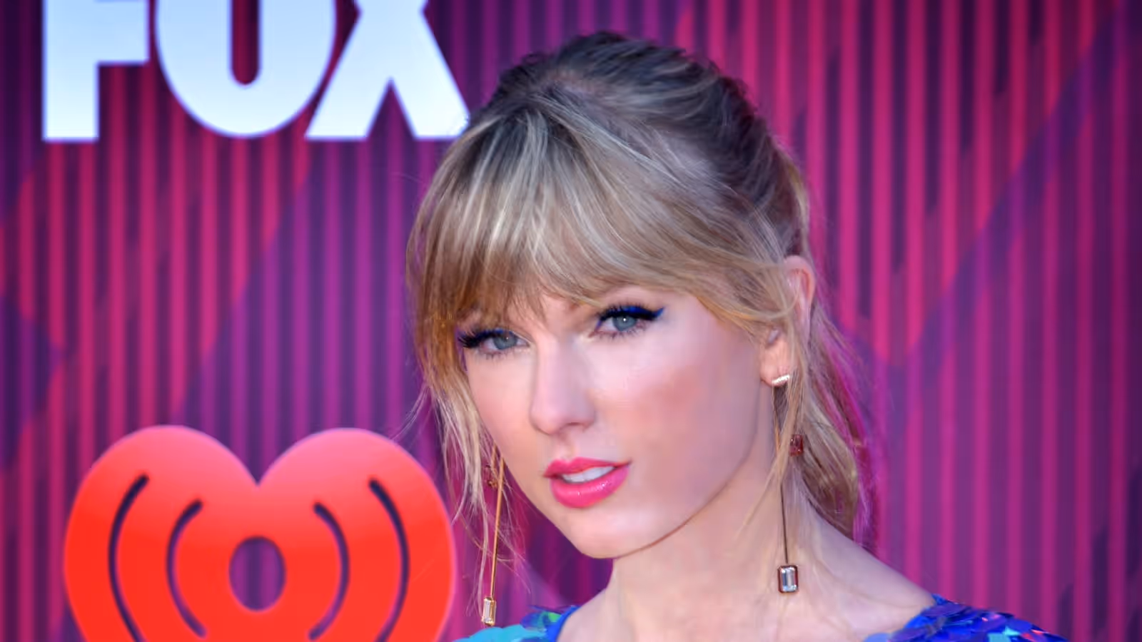 Taylor Swift facing the camera in a head-and-shoulders portrait, blonde hair up with a fringe, gold drop earrings and a blue sequinned top, in front of a magenta backdrop carrying the FOX and iHeartRadio logos.