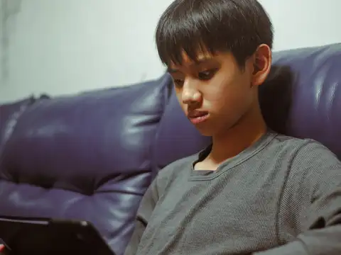 A boy sitting back on a sofa, looking down at a tablet held in his lap.