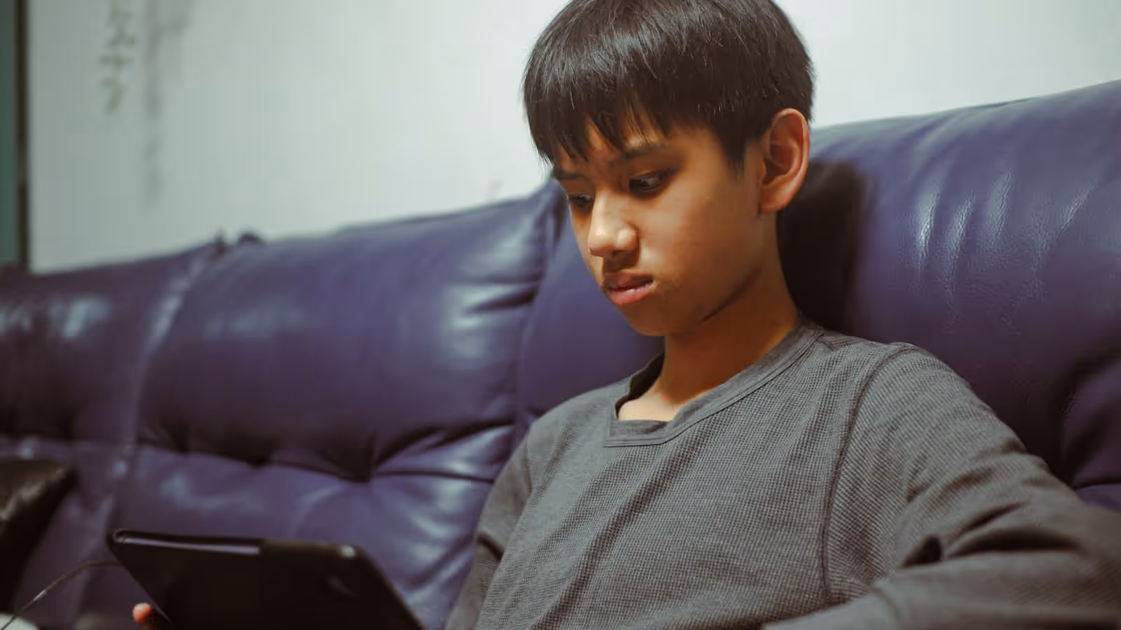 A boy sitting back on a sofa, looking down at a tablet held in his lap.