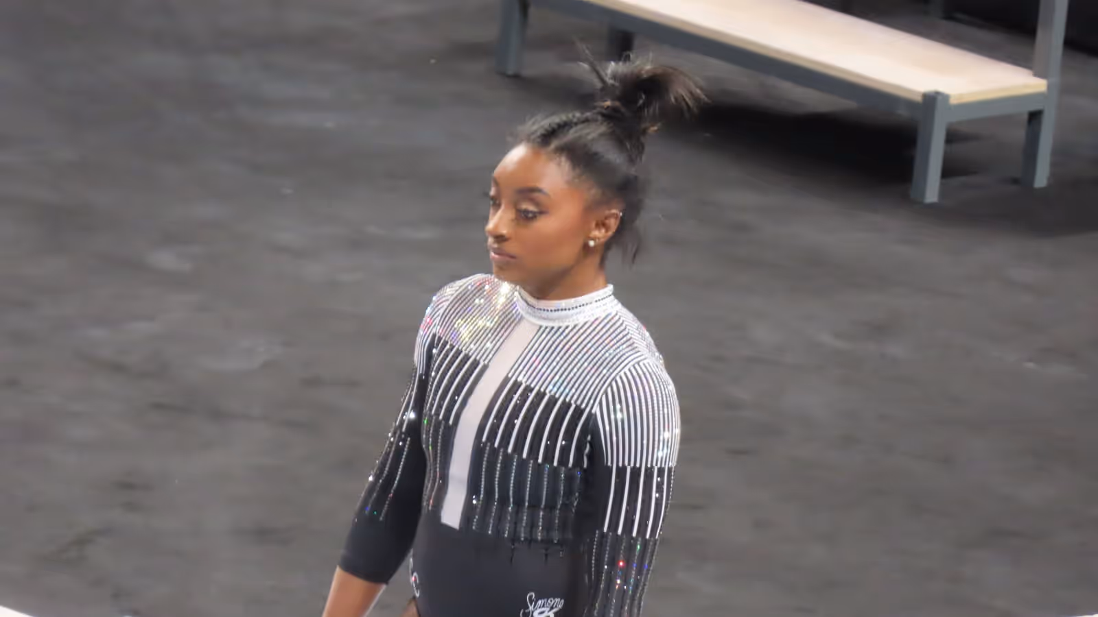 Simone Biles standing on a competition floor in a black and silver sparkling leotard, hair tied up, looking to one side, with a bench and grey matting behind her.