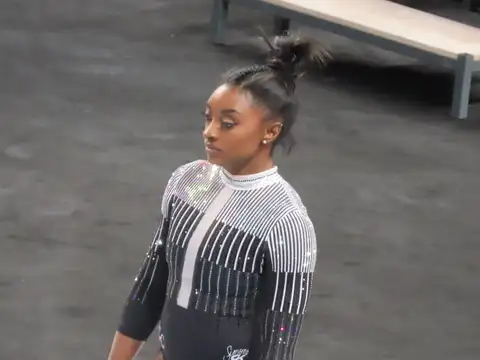 Simone Biles standing on a competition floor in a black and silver sparkling leotard, hair tied up, looking to one side, with a bench and grey matting behind her.