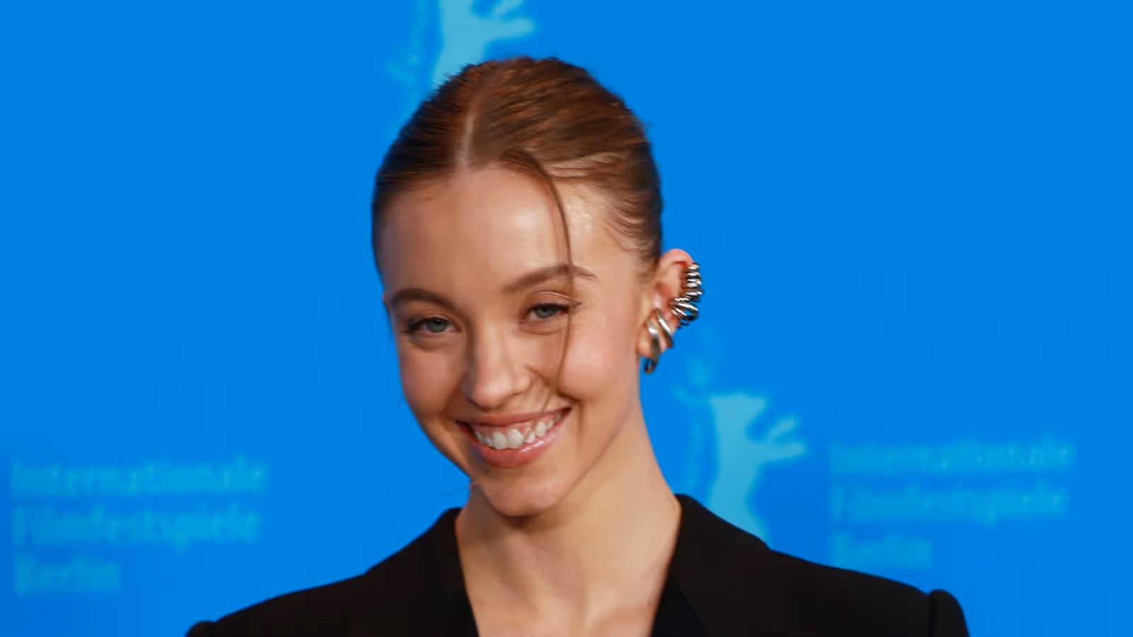 Sydney Sweeney em uma fotografia de cabeça e ombros, sorrindo, cabelo preso, vestindo uma jaqueta preta e um brinco de orelha prateado, em frente a um fundo azul do Berlinale.