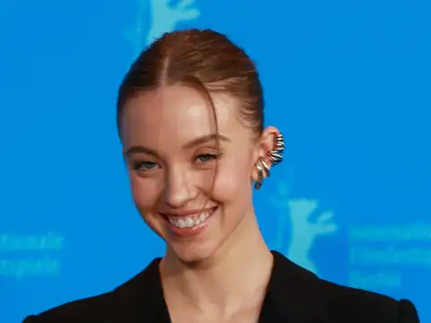 Sydney Sweeney in a head-and-shoulders photograph, smiling, hair tied back, wearing a black jacket and a silver ear cuff, in front of a blue Berlinale backdrop.