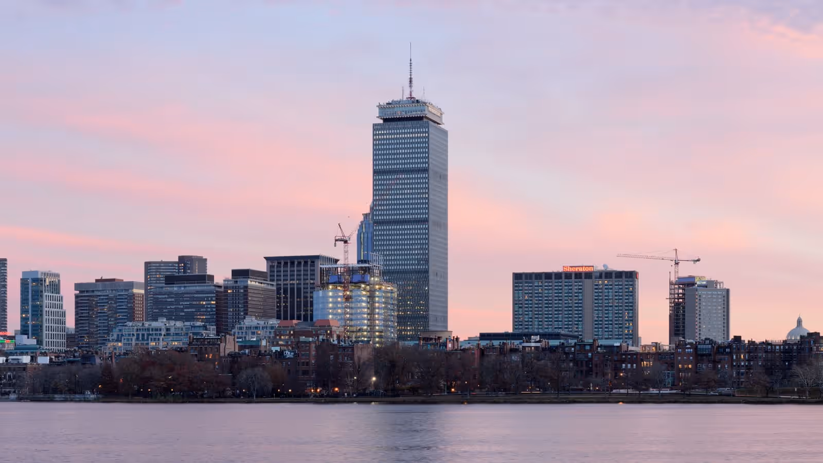 Die Skyline von Boston bei Dämmerung, gesehen über den Charles River, mit dem Prudential Tower, der über Back Bay unter einem rosa Himmel emporragt.