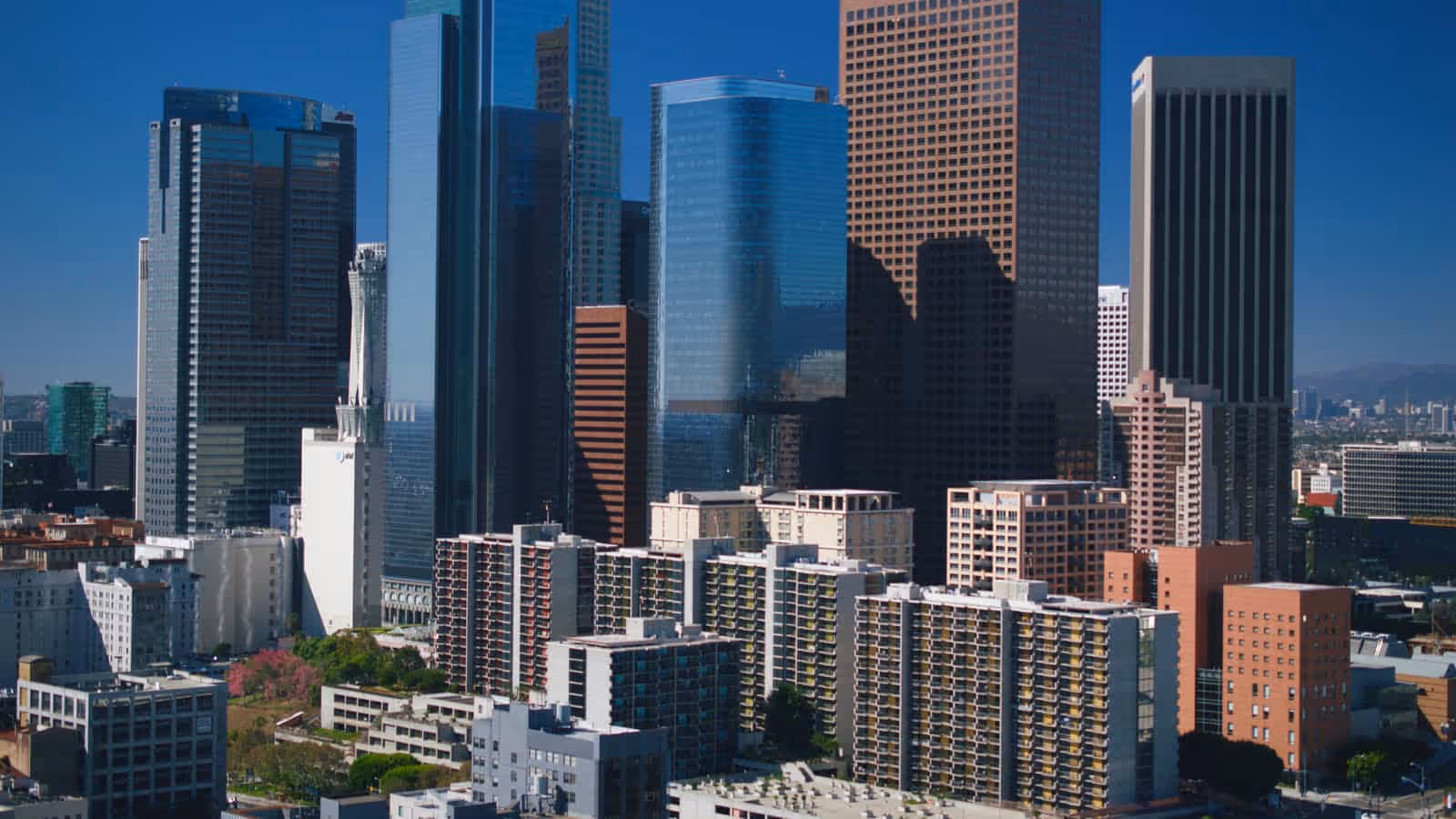 Downtown Los Angeles high-rise towers photographed from above in bright daylight, with lower apartment blocks in the foreground and hills on the horizon.