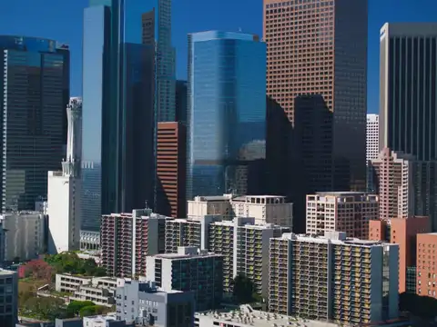 Downtown Los Angeles high-rise towers photographed from above in bright daylight, with lower apartment blocks in the foreground and hills on the horizon.