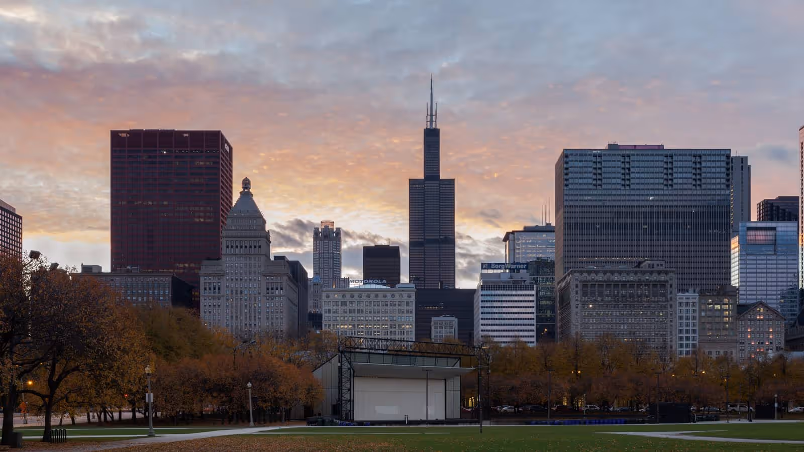 El horizonte de Chicago al anochecer visto desde el Parque Grant, con la Torre Willis y sus antenas en el centro y árboles de otoño en primer plano.
