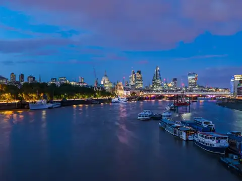 The River Thames at dusk seen from a bridge, with the dome of St Paul’s Cathedral and the lit towers of the City of London along the far bank, a road bridge crossing on the right and moored boats in the foreground.