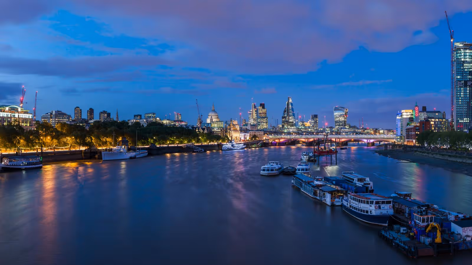 The River Thames at dusk seen from a bridge, with the dome of St Paul’s Cathedral and the lit towers of the City of London along the far bank, a road bridge crossing on the right and moored boats in the foreground.