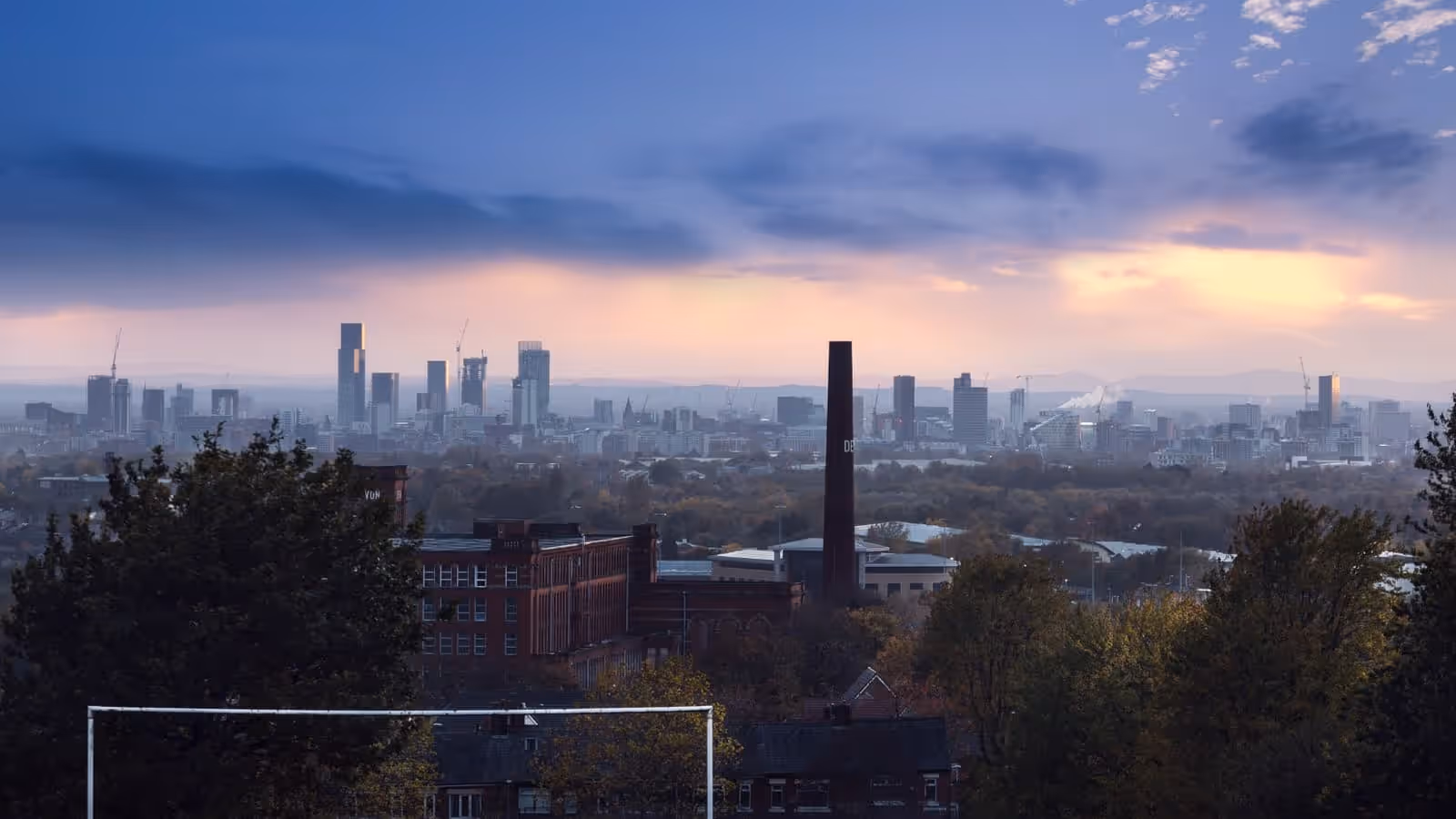 The Manchester skyline at dusk seen from high ground to the east, with the city centre towers on the horizon and a red-brick mill and chimney in the foreground.