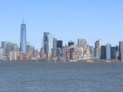 The Lower Manhattan skyline photographed across open water on a clear day, with One World Trade Center rising at the left of the cluster of towers.