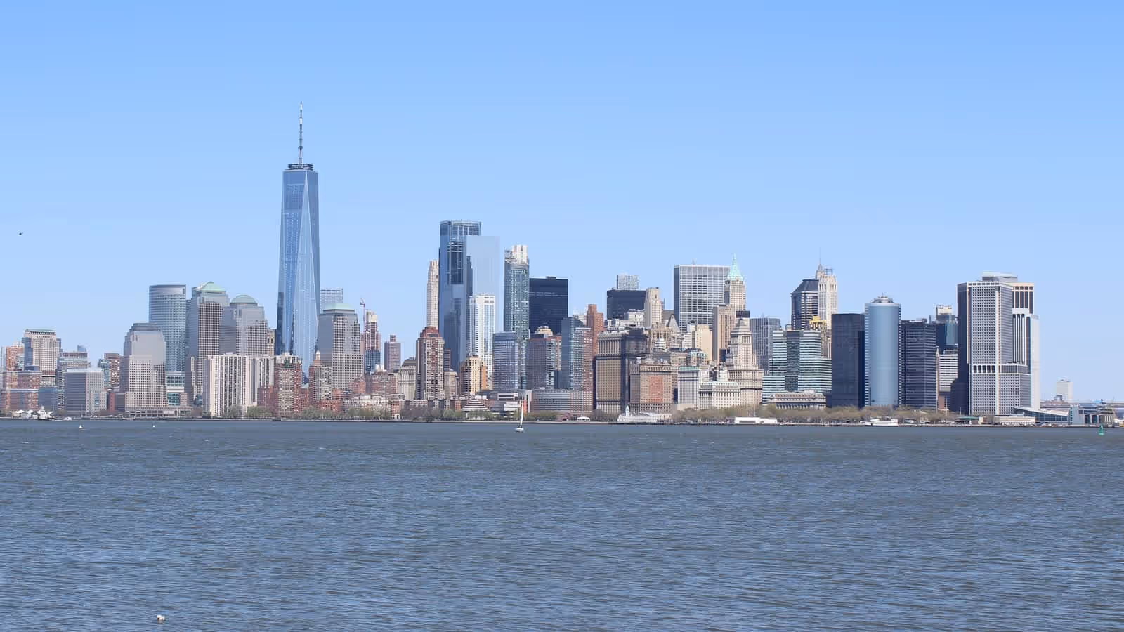 The Lower Manhattan skyline photographed across open water on a clear day, with One World Trade Center rising at the left of the cluster of towers.