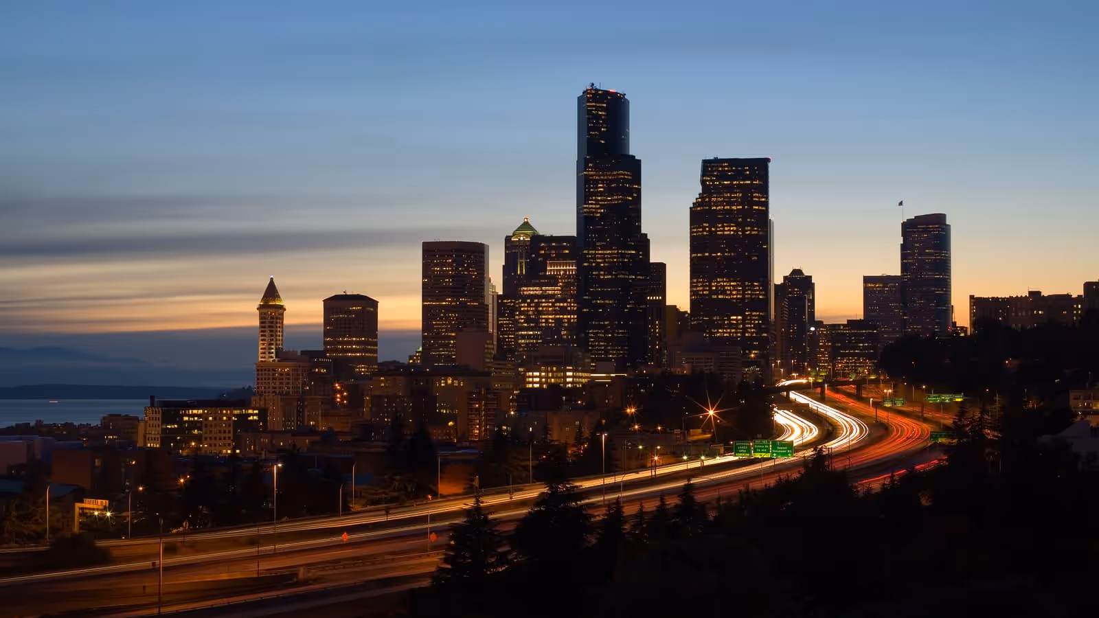 The downtown Seattle skyline at dusk, with the Columbia Center and the pyramid-topped Smith Tower above Interstate 5, light trails from traffic below and Elliott Bay beyond.